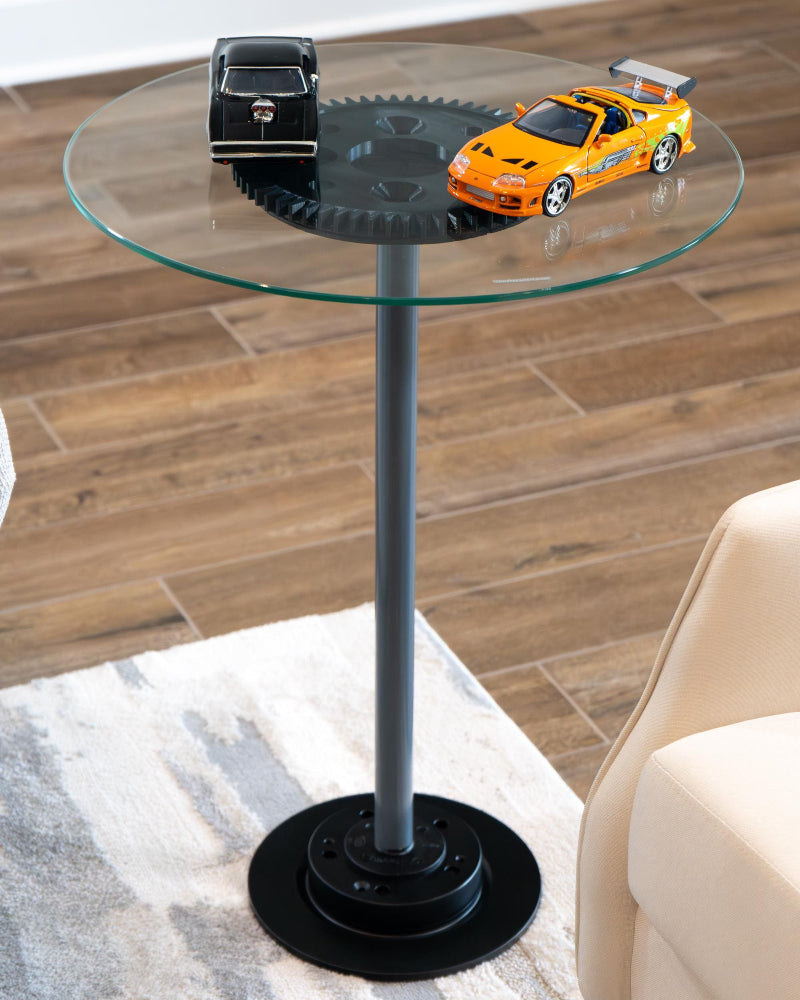 Glass side table with black base featuring model cars on a wooden floor.