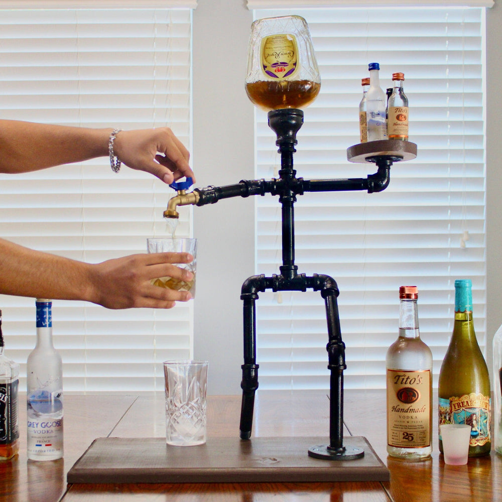 Person pouring a drink from a tap into a glass, with various bottles on a table.