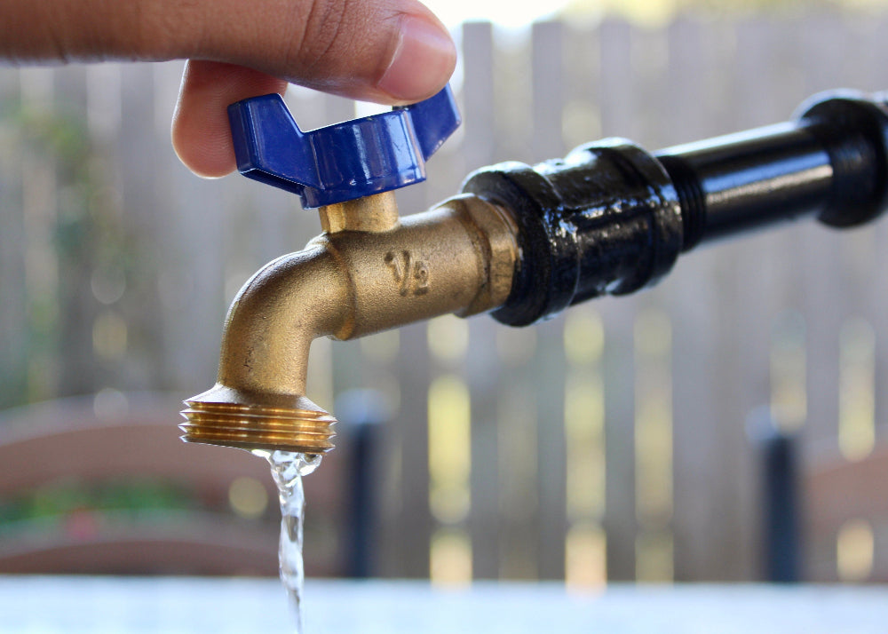Brass faucet with blue handle being turned on, water flowing, outdoor setting.