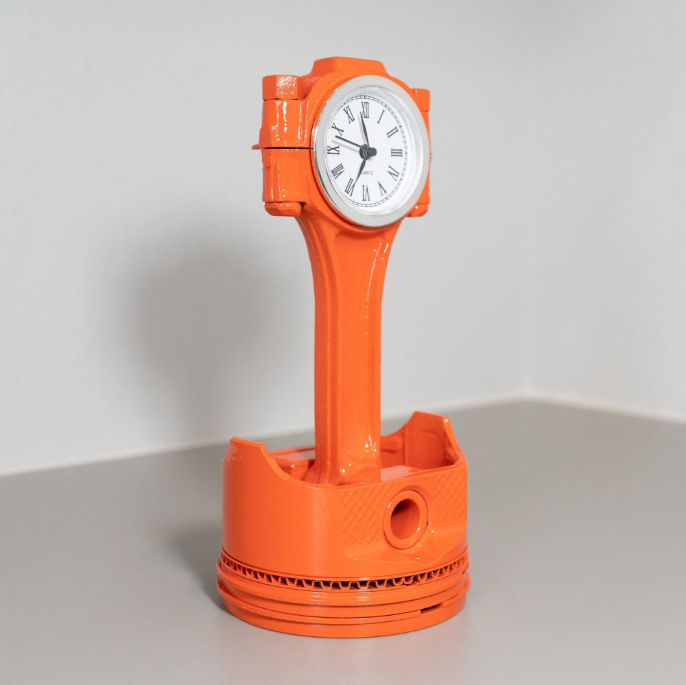 Orange clock shaped like a piston on a gray surface with a white background