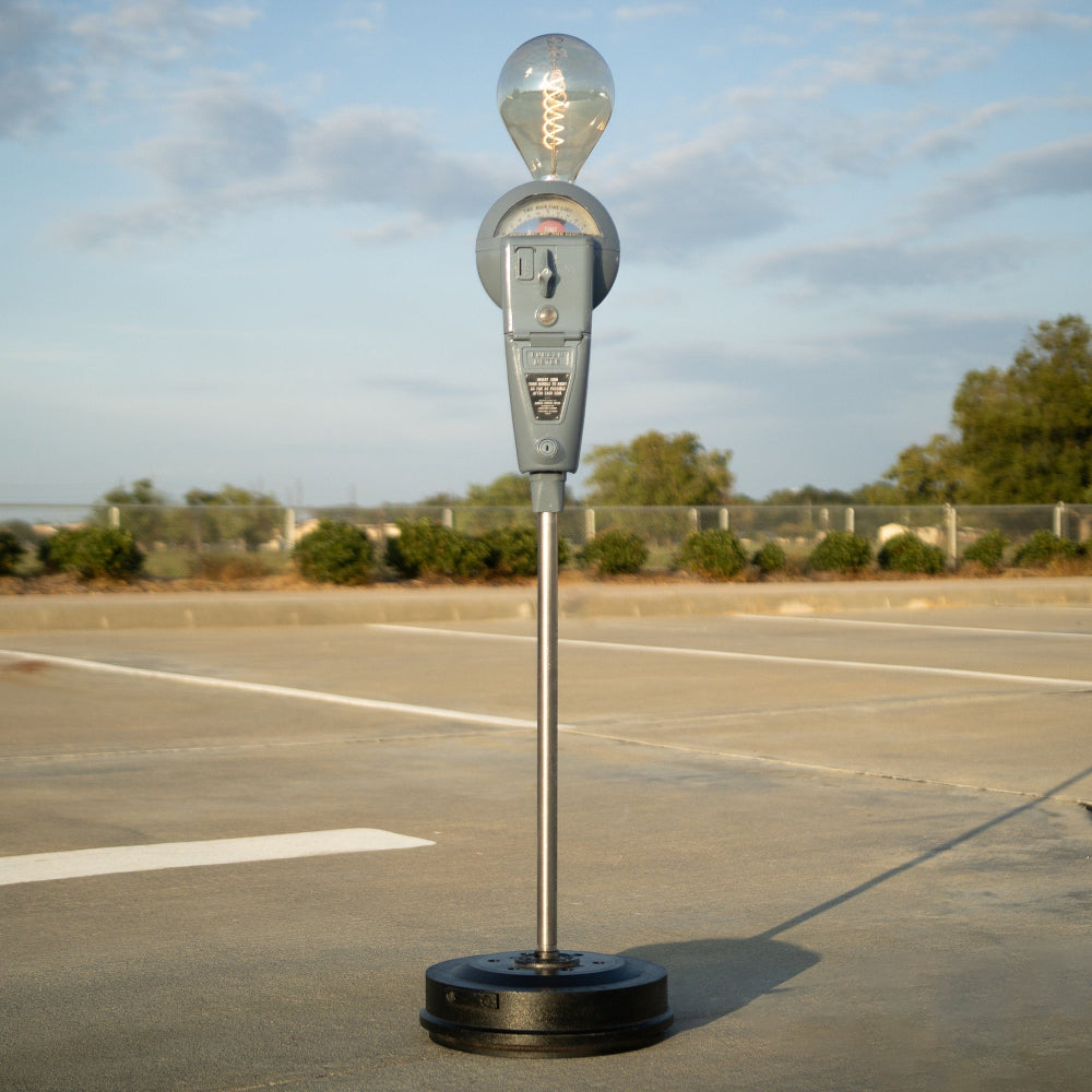 Decorative light fixture shaped like a parking meter on a concrete surface with trees in the background.