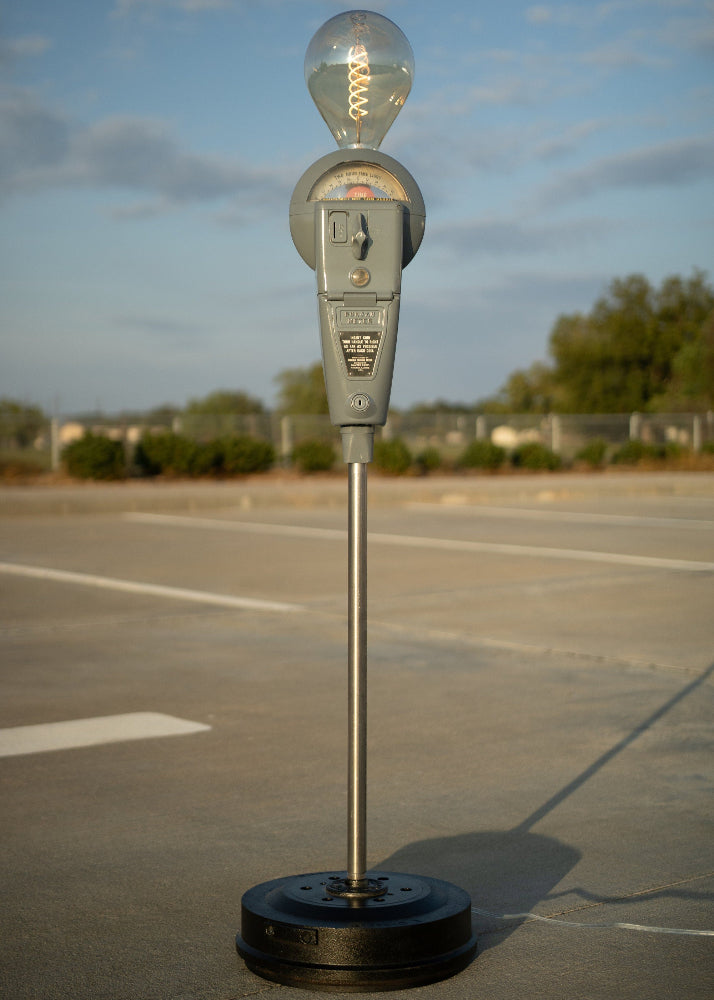 Parking meter with a light bulb on top in an open area with trees and sky.