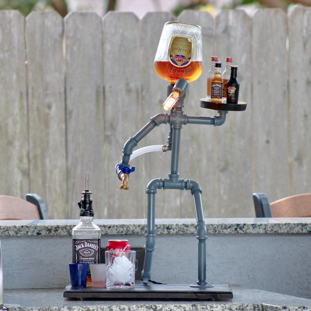 Pipe sculpture with a face, holding a glass of beer and surrounded by bottles on a patio.