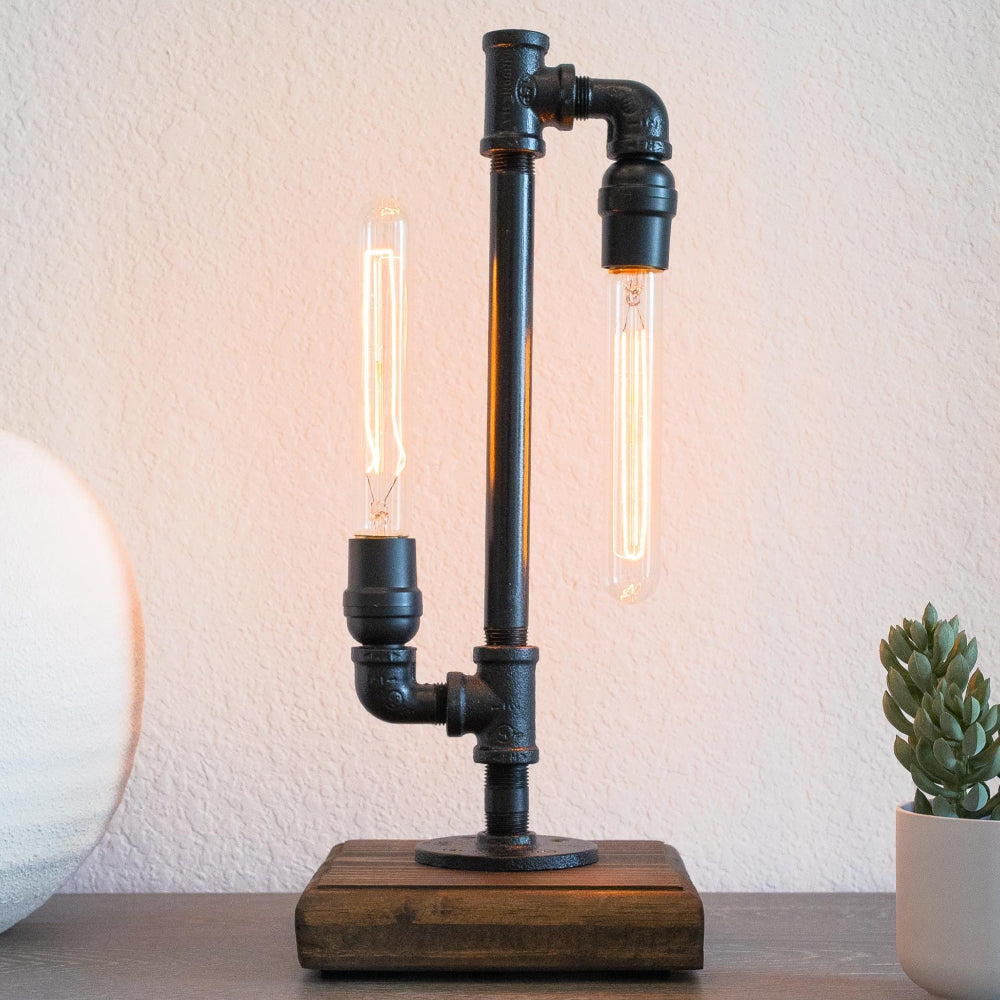 Table lamp with industrial design featuring pipes and wooden base, illuminated with two visible light bulbs, on a surface with a white vase and small plant in the background.