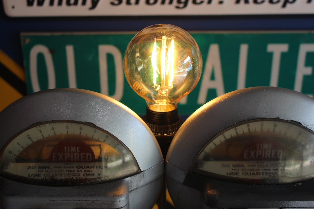 Vintage-style light bulb between two old parking meters with a sign in the background.
