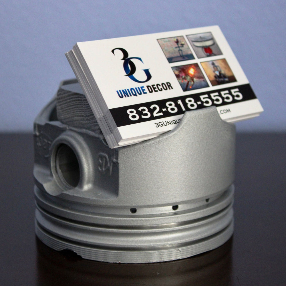 Metallic engine piston with a business card holder displaying 'Unique Decor' contact information.