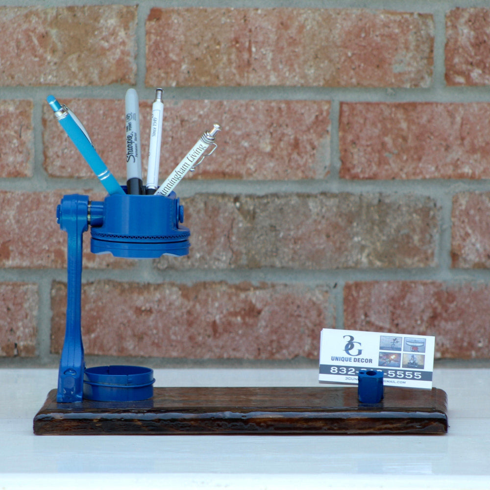 Blue pen holder with pens on a wooden base against a brick wall.