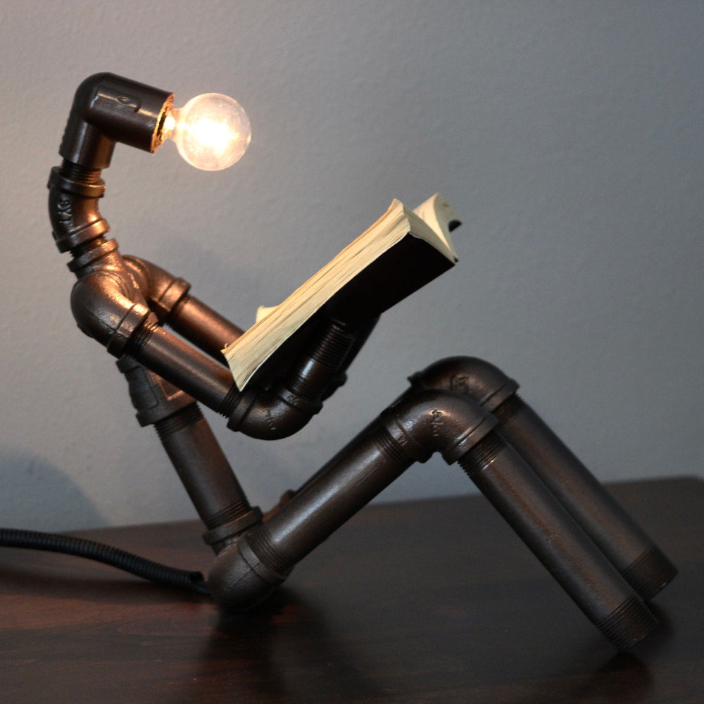 Lamp made from pipes with a light bulb, resembling a person reading a book.