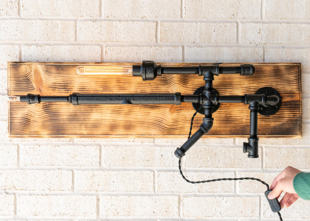 Wall-mounted industrial-style light fixture with wooden panel and black pipes on a white brick wall.