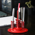 Red pen holder with multiple pens on a desk with a laptop in the background
