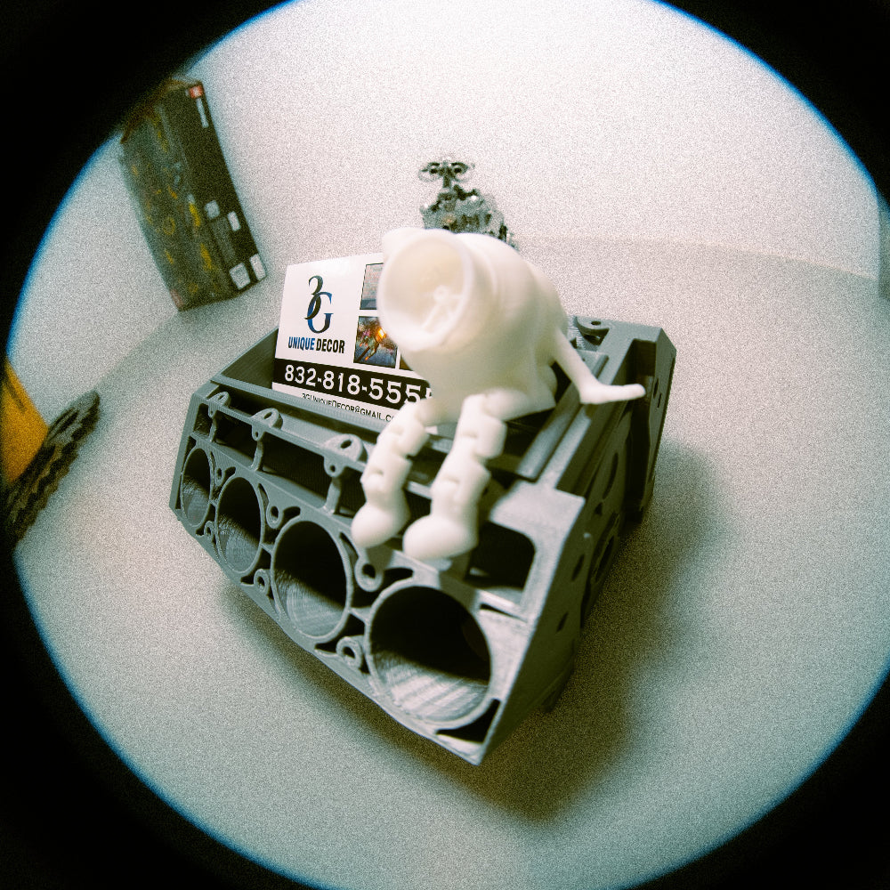 Small white robot sitting on a mechanical engine block with a business card on a light surface.