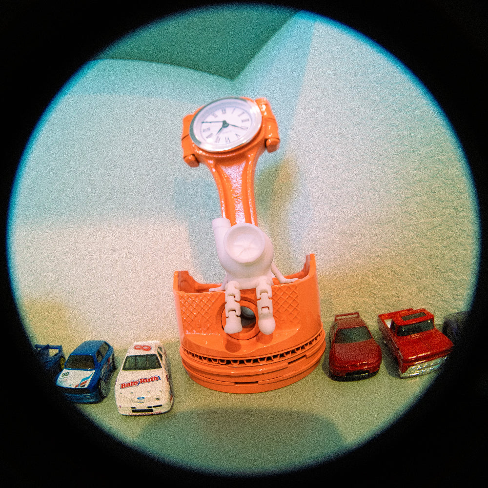 Orange toy car with a clock face and small toy cars on a textured surface
