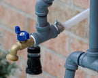 Outdoor faucet with blue handle attached to gray pipes against a brick wall.
