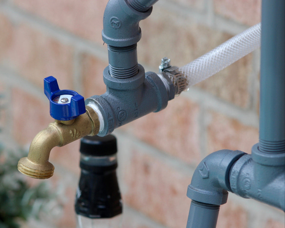 Outdoor faucet with blue handle attached to gray pipes against a brick wall.