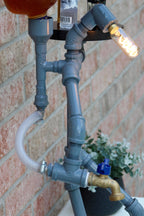Upcycled lamp made from gray pipes and fittings against a brick wall.