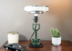 Table lamp with vintage design on a wooden surface, next to a small model car and a potted plant.