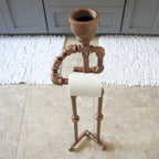 Bathroom setting with a decorative toilet paper holder made of brown pipes on a white bath mat.