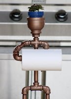 Pipe-themed toilet paper holder with a small potted plant on top against a metallic background.