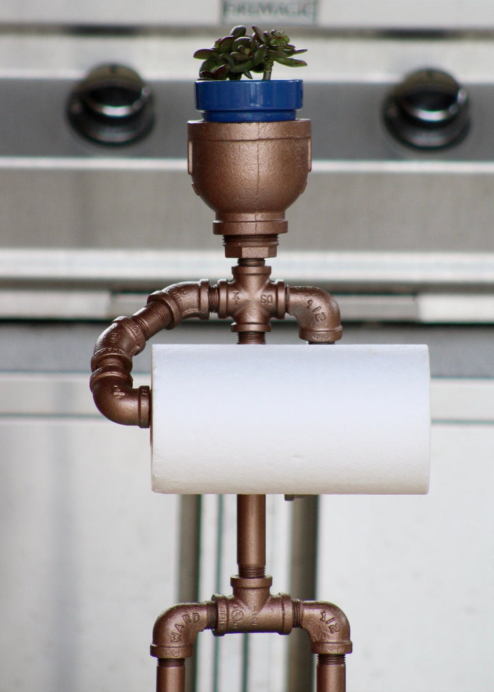 Pipe-themed toilet paper holder with a small potted plant on top against a metallic background.