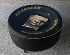 Round black tire-shaped ottoman with 'Wrangler' branding on a gray floor.