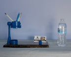 Blue mechanical desk organizer with pens and a bottle of water on a wooden surface against a blue wall.