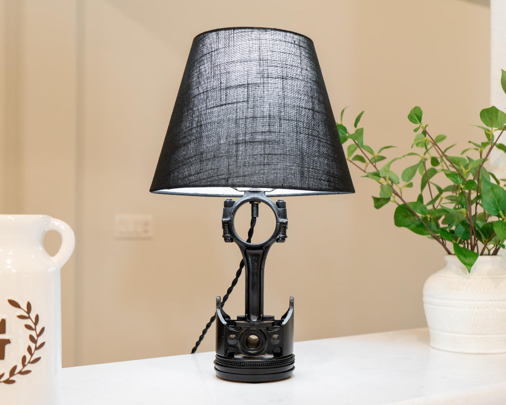 Black table lamp with a textured shade on a white surface, with a plant and decorative item in the background.