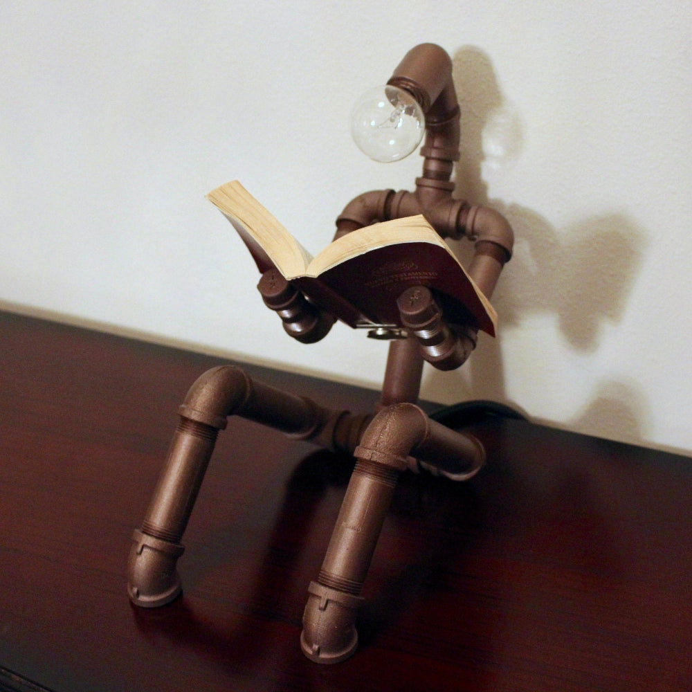 Pipe sculpture with a light bulb head reading a book on a wooden surface.