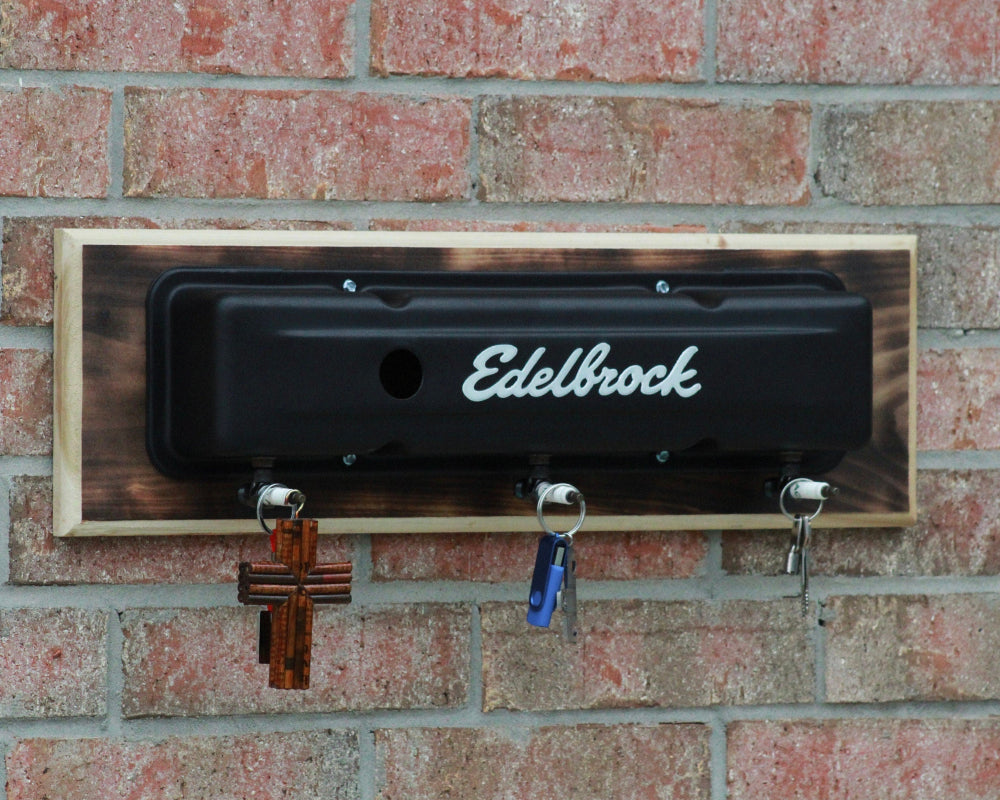 Black Edelbrock engine cover mounted on a brick wall with hooks attached.